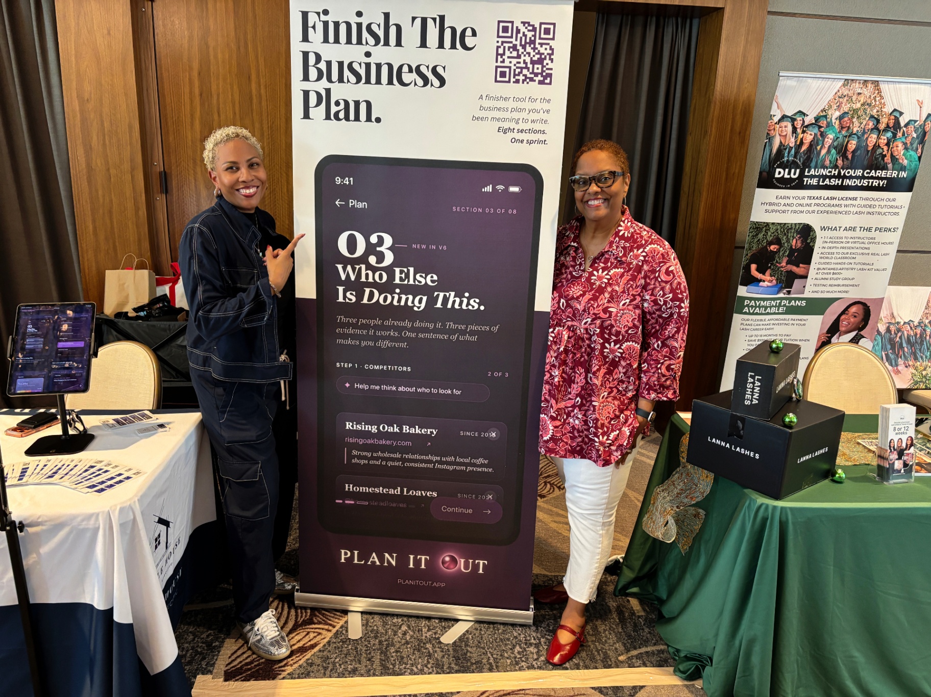 Two people standing beside the Finish The Business Plan banner at the Chronicle House booth.